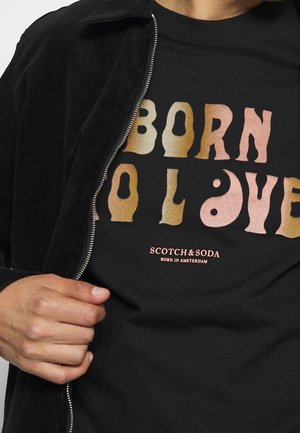 Personne portant un t-shirt noir avec "BORN TO LOVE" en lettres ondulées et multicolores et le texte "SCOTCH & SODA BORN IN AMSTERDAM" en dessous.