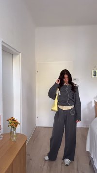 Maglione grigio oversize, camicia button-down crema, pantaloni grigi a gamba larga, borsa gialla, sneakers bianche; interno minimalista con mobili in legno.