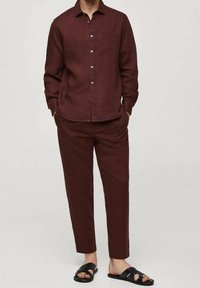 Mann in einem bordeauxroten Hemd mit Knöpfen und passenden Hosen, der mit den Händen in den Taschen vor einem schlichten Hintergrund steht. Er trägt schwarze Sandalen mit offenen Zehen.