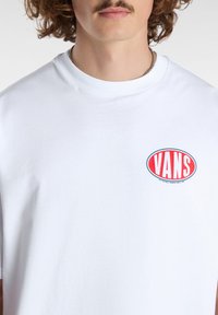 Bílá bavlněná tričko s červeným a modrým oválným logem Vans na levé části hrudi. Klasický design s kulatým výstřihem, krátké rukávy a hladká textura.