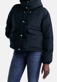 Svart vadderad jacka med huva, med tryckknappar, två framfickor och elastiska manschetter. Slät tyg med sydda detaljer.