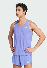 Hombre con camiseta de tirantes atlética morada y pantalones cortos con pequeñas perforaciones y logotipos amarillos de Adidas, de pie con las manos en las caderas, mirando hacia la izquierda.