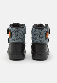 Baskets montantes noires et teal avec un motif ludique, des bandes Velcro et un matériau texturé. Caractérisées par un logo circulaire en orange.