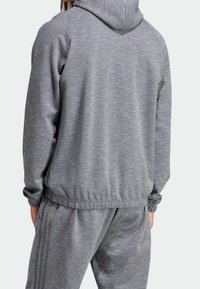 Person i grå huvtröja och matchande joggingsbyxor med tre mörkare grå ränder på sidan, sedd bakifrån.