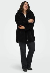 Melna kapuce frotē jaka ar mīkstu, teksturētu virsmu, priekšējo rāvējslēdzēju, sānu kabatām un relaksētu piegriezumu; valkāta ar tumšām džinsām.