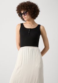 Top de tirantes negro con textura acanalada y tres botones de metal en la parte delantera, combinado con una falda blanca fluida hecha de un tejido ligero.