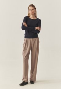 Laivastonsininen neulottu pusero pyöreällä kaula-aukolla, yhdistetty vaaleanruskeisiin laskostettuihin housuihin ja tummiin kenkiin, neutraalia taustaa vasten.