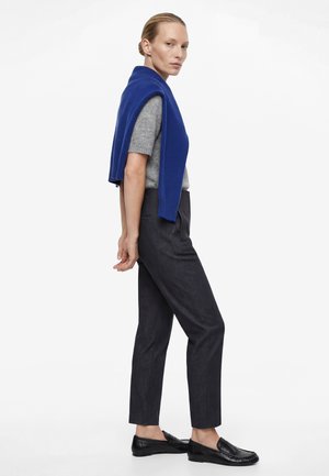 Frau mit blondem Haar in einem grauen Kurzarmpullover, dunkelblauer Hose, schwarzen Slippern und einem blauen Pullover über den Schultern stehend, seitlich gesehen.