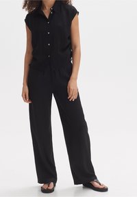 Černá košile s krátkými rukávy, límečkem a knoflíky, doplněná o černé kalhoty s širokými nohavicemi. Hladká textura, volný střih, minimální počet kovových doplňků.