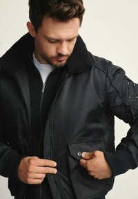 Mężczyzna w czarnej kurtce bomberce z futrzanym kołnierzem i ściągaczami, zapinający guzik w przedniej kieszeni, patrząc w dół.