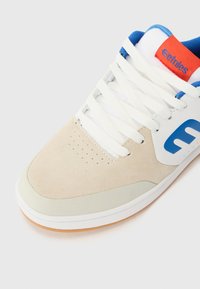 Sapatilha de camurça bege com detalhes em pele branca e pormenores azuis. Apresenta atacadores brancos e uma etiqueta vermelha com o logótipo "etnies". Solado de borracha plano.