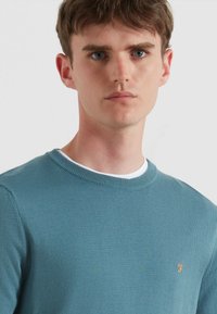 Niebieski sweter z okrągłym dekoltem, z prążkowanym kołnierzem, wykonany z teksturowanej dzianiny, z małym haftowanym pomarańczowym logo na piersi.