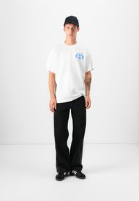 White cotton t-shirt with a blue graphic design on the left chest, paired with loose-fitting black denim jeans and black sneakers with white stripes.