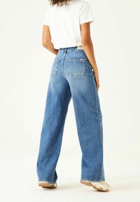 Jeans de denim de pierna ancha en azul medio, de cintura alta con un diseño de cinco bolsillos. Presentan costuras resistentes y un acabado de dobladillo sin rematar.