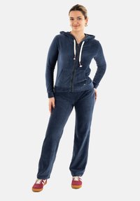 Chándal de terciopelo acanalado en navy con una sudadera con cremallera, bolsillos delanteros y cordones en contraste; combinado con pantalones a juego y zapatillas en color burdeos.