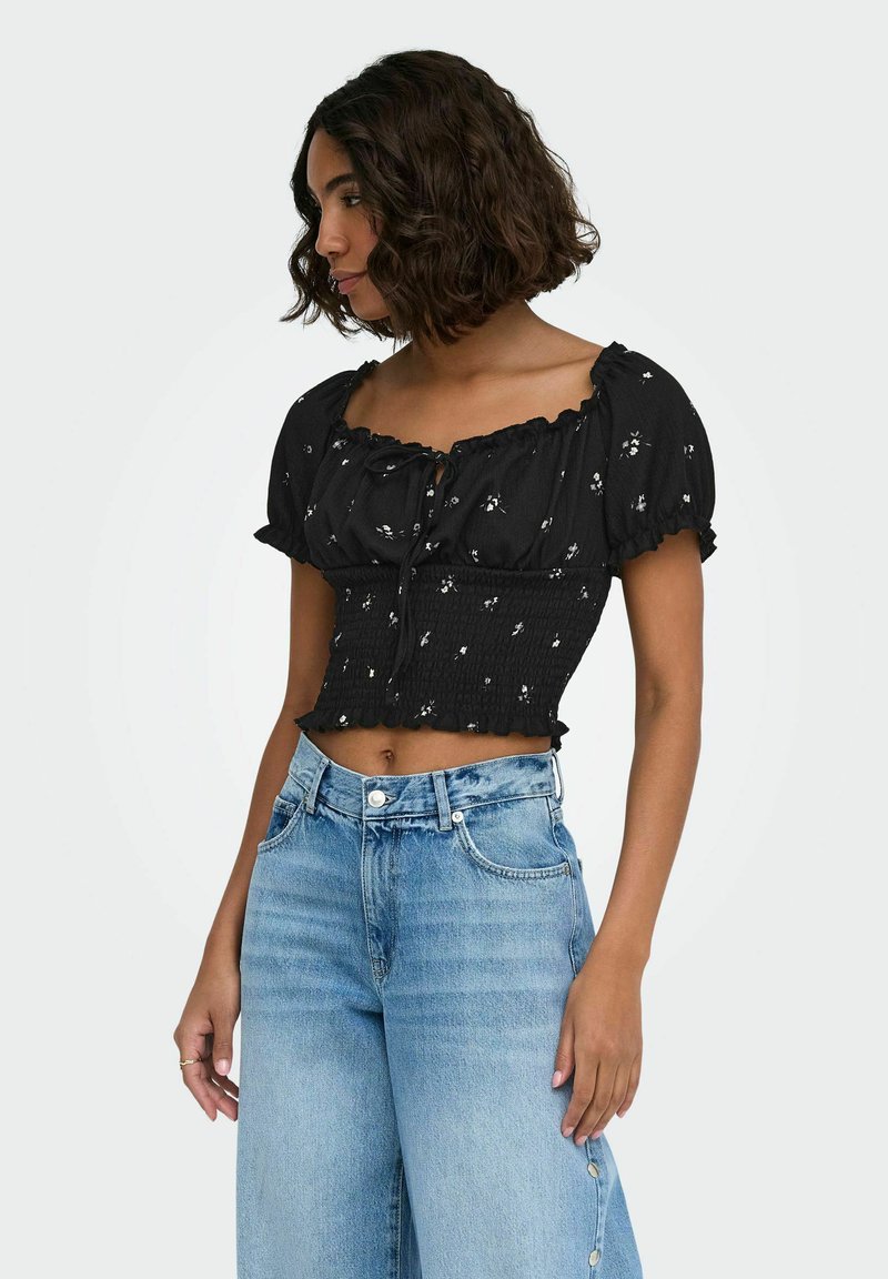 Jeune femme aux cheveux bouclés portant un chemisier court noir à manches courtes avec de petites fleurs blanches et un jean taille haute bleu clair.