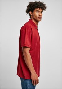 Rød kortermet poloskjorte med krage, laget av glatt stoff. Foran kanten har den sidesslister; brukt med blå jeans.