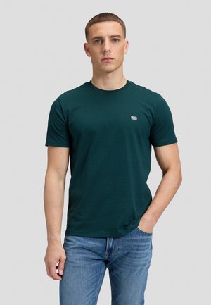Homme portant un t-shirt Lee vert foncé et un jean bleu, debout avec une main dans la poche devant un fond uni.