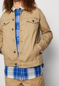 Burgundowa kurtka dżinsowa z zapięciem na guziki i dwiema kieszeniami na piersi, wyłożona na niebieskiej i białej koszuli w kratę. Widoczna tekstura materiału.