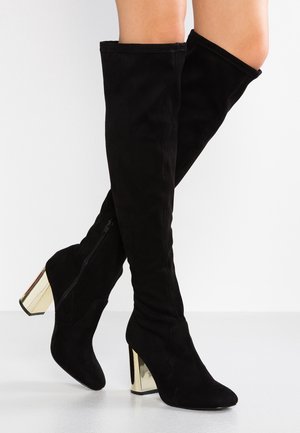 Musta mokkanahkainen polven yli ulottuva saappaat, joissa on kullanväriset paksut korot, jalat ristissä, näkyvissä sivuvetoketju yhdessä saappaassa.