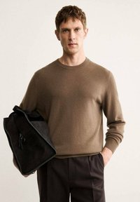 Brauner gestrickter Pullover mit Rundhalsausschnitt, getragen mit gefalteten dunklen Hosen. Hält eine schwarze Wildledertasche mit Reißverschluss und Quaste.