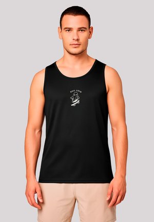 Young man wearing a black tank top with a white ghost on skateboard graphic and beige shorts, standing against a plain light background.