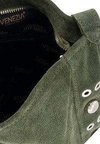 Grøn suedepung med lynlåslukning, runde metalliske accenter og synlige sting. Indersiden er foret med mønstret stof.