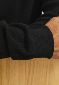 Fekete kötött pulóver ujja, amely puha, texturált felülettel és bordázott részletekkel rendelkezik, fa felületen.