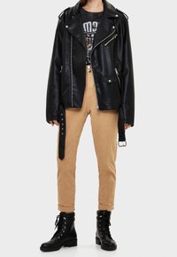 Chaqueta de moto de piel sintética negra con herrajes plateados, pantalones beiges y botas negras con cordones. La chaqueta cuenta con cremalleras y charreteras en los hombros.