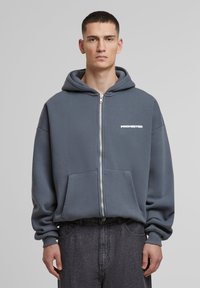 Gray zip-up hoodie in soft fabric with front pockets and white "PROHIBITED" text. Features a drawstring hood and ribbed cuffs.