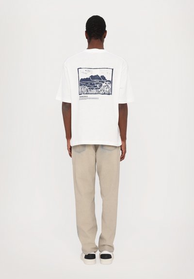 Tricou alb din bumbac cu un grafic albastru pe spate care ilustrează munți și apă, asortat cu pantaloni bej și încălțăminte cu accente negre.