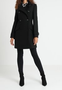 Svart dubbelknäppt rock med skräddarsydd passform, utrustad med sidofickor och knappdetaljer, kombinerad med svarta slim-fit jeans och ankelstövlar.