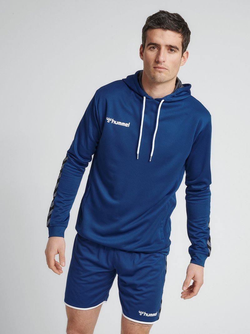Mand iført blå Hummel sportshoodie med hvide snorer og matchende shorts, stående mod en ensfarvet lys baggrund.
