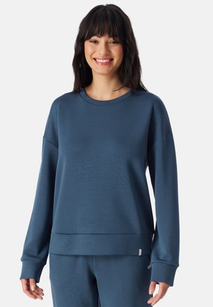 Ung kvinde med mørkt hår smiler, iført en løs mørkeblå langærmet sweatshirt og matchende bukser mod en ensfarvet baggrund.