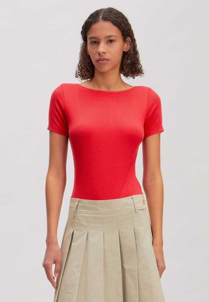 Vrouw met krullend haar draagt een aansluitend rood topje met korte mouwen en een beige geplooide rok, staand tegen een egale lichte achtergrond.