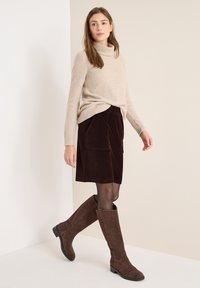 Sweter z wysokim kołnierzem w kolorze kremowym, brązowa spódnica z przędzy kordowej z kieszeniami oraz brązowe zamszowe botki za kolano na niskim obcasie. Gładka faktura.