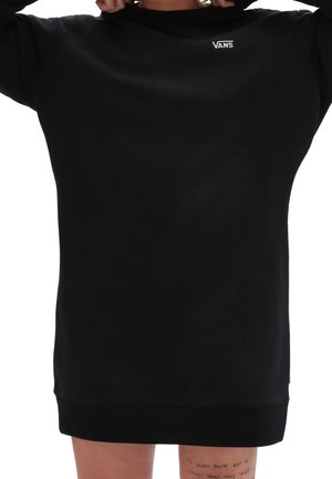 Persona che indossa un vestito felpa nero Vans, con le mani che tengono il colletto, con un tatuaggio testuale visibile sulla coscia superiore.