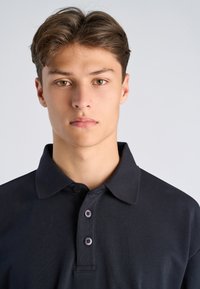 Jonge man met kort bruin haar en hazelnootkleurige ogen, gekleed in een zwart polohemd met knopen, tegen een effen lichtgrijze achtergrond.