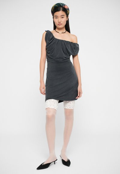 Vestido cinza drapeado de um ombro com textura suave, combinado com calções de renda brancos e meias transparentes. Sapatos de salto agulha pretos completam o visual.