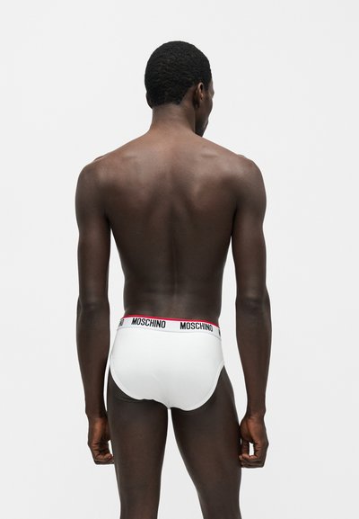 Ανδρικό μοντέλο με σκουρόχρωμο δέρμα και κοντά μαλλιά, εικονιζόμενο από πίσω, φορά λευκά εσώρουχα με λωρίδα μέσης με το εμπορικό σήμα "Moschino".