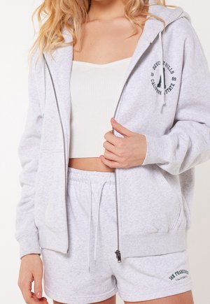 Hoodie zippé gris clair avec poches avant, arborant un logo noir imprimé. Associé à un short gris clair assorti avec un détail brodé.