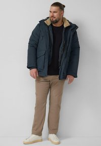 Gewatteerde marineblauwe winterparka met met bont gevoerde capuchon, ritssluiting, meerdere voorzakken, gecombineerd met beige broek en witte sneakers.