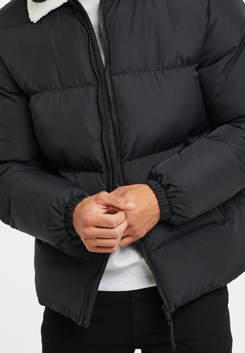 Homme portant une veste matelassée noire avec des poignets élastiques et une chemise blanche en dessous, mains jointes à la taille, veste partiellement zippée.