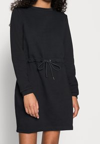 Svart långärmad casual klänning med rund halsringning och dragsko i midjan. Tillverkad av mjukt tyg, med en avslappnad passform och minimala detaljer.