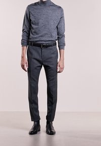 Hombre vistiendo un suéter gris de manga larga, pantalones ajustados gris oscuro con un cinturón negro y botas negras por el tobillo, de pie sobre un fondo liso.