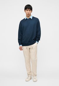 Ναυτικό πουλόβερ με στρογγυλή λαιμόκοψη, φορεμένο πάνω από ένα ανοιχτό γαλάζιο πουκάμισο με γιακά. Ανοιχτό μπεζ παντελόνι με κορδόνι στη μέση, συνδυασμένο με λευκά αθλητικά παπούτσια.