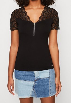 Zwart kort mouwtjes shirt met kanten schouders en een ritssluiting aan de voorkant. Gecombineerd met een spijkerbroek minirok in patchwork-stijl met blauwtinten.