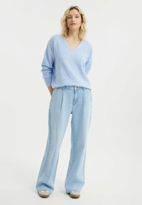 Hellblauer Strickpullover mit V-Ausschnitt, kombiniert mit hellblauen weiten Jeans und beigen Turnschuhen. Weiche Textur und lässige Passform.