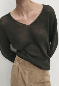 Ciemnozielony sweter dżersejowy z dekoltem w kształcie V, luźny krój i teksturowana tkanina, zestawiony z jasnobeżowymi spodniami z zamszu.