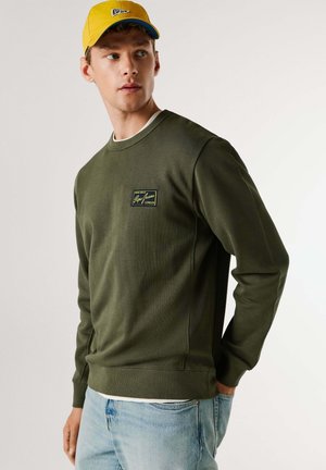 Alyvuogių žalumos džemperis su gofruotais rankogaliais ir apačia, ant krūtinės yra mažas geltonos ir mėlynos spalvų firminis lopas. Dėvimas su šviesiai mėlynomis džinsinėmis kelnėmis.